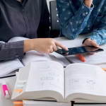 Two people using a tablet on a table covered in books and pens