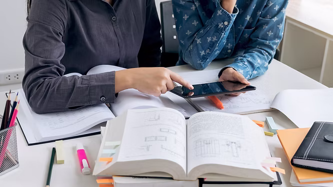 Two people using a tablet on a table covered in books and pens