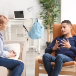 A young man sitting in an orange armchair speaking to a therapist