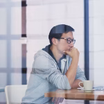 A man with glasses sitting at a wooden table looking at his laptop